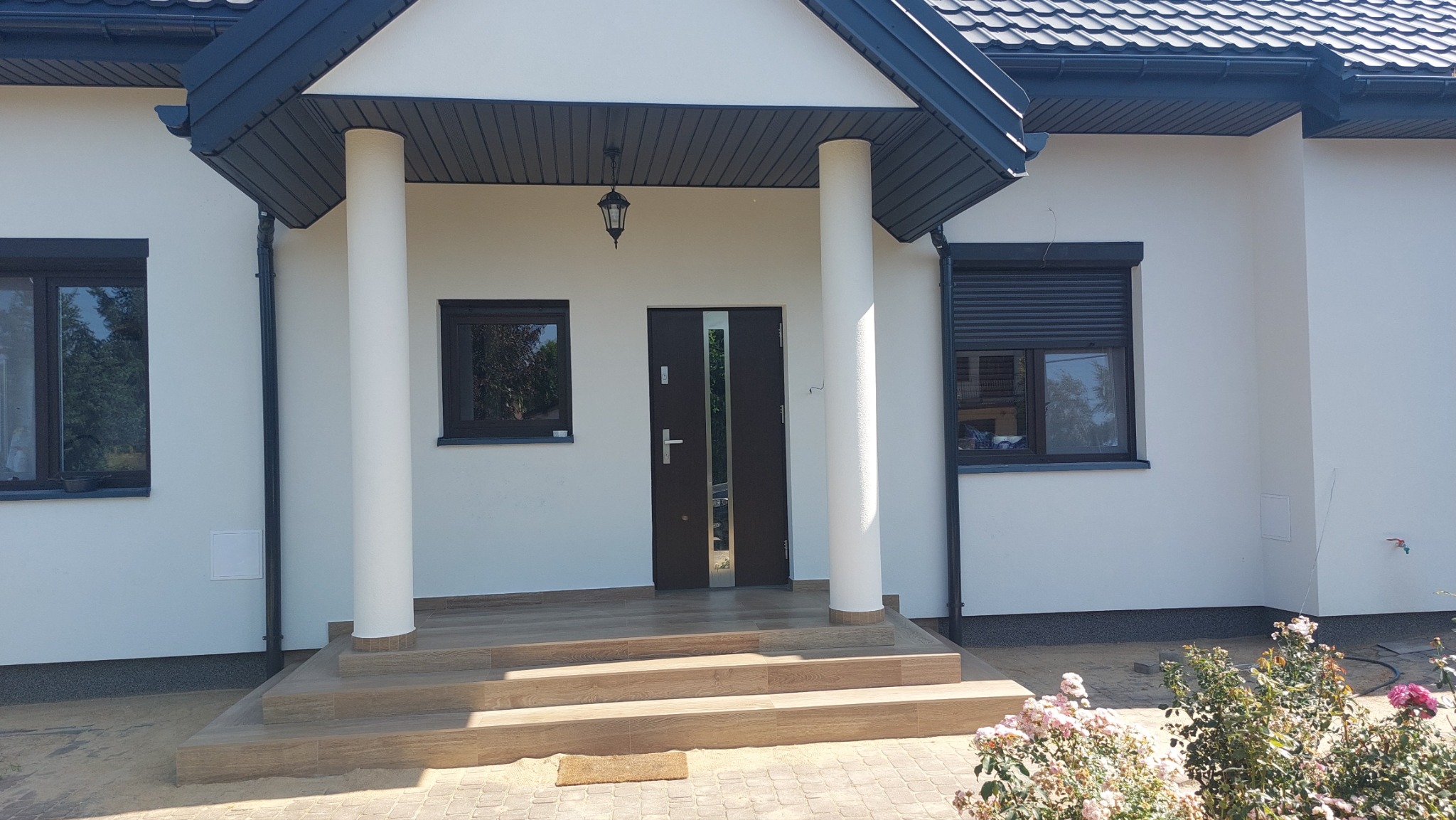 Wykończona elewacja domu jednorodzinnego w Jaktorowie z kolumnami, schodami wejściowymi wyłożonymi płytkami imitującymi drewno, ciemnymi oknami z roletami i nowoczesnymi drzwiami wejściowymi.