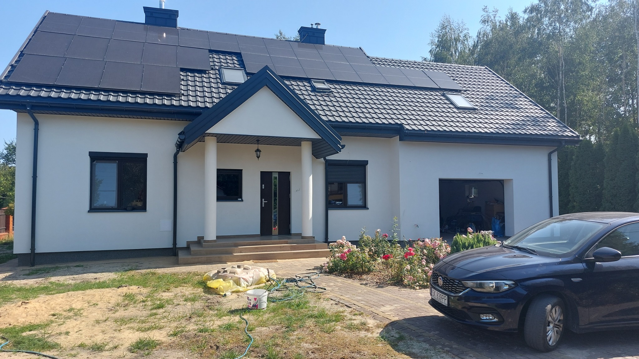 Biały dom jednorodzinny z ciemnymi panelami słonecznymi na dachu, ciemnymi oknami z roletami i podjazdem z kostki brukowej, z zaparkowanym ciemnym samochodem osobowym i workami z materiałem...