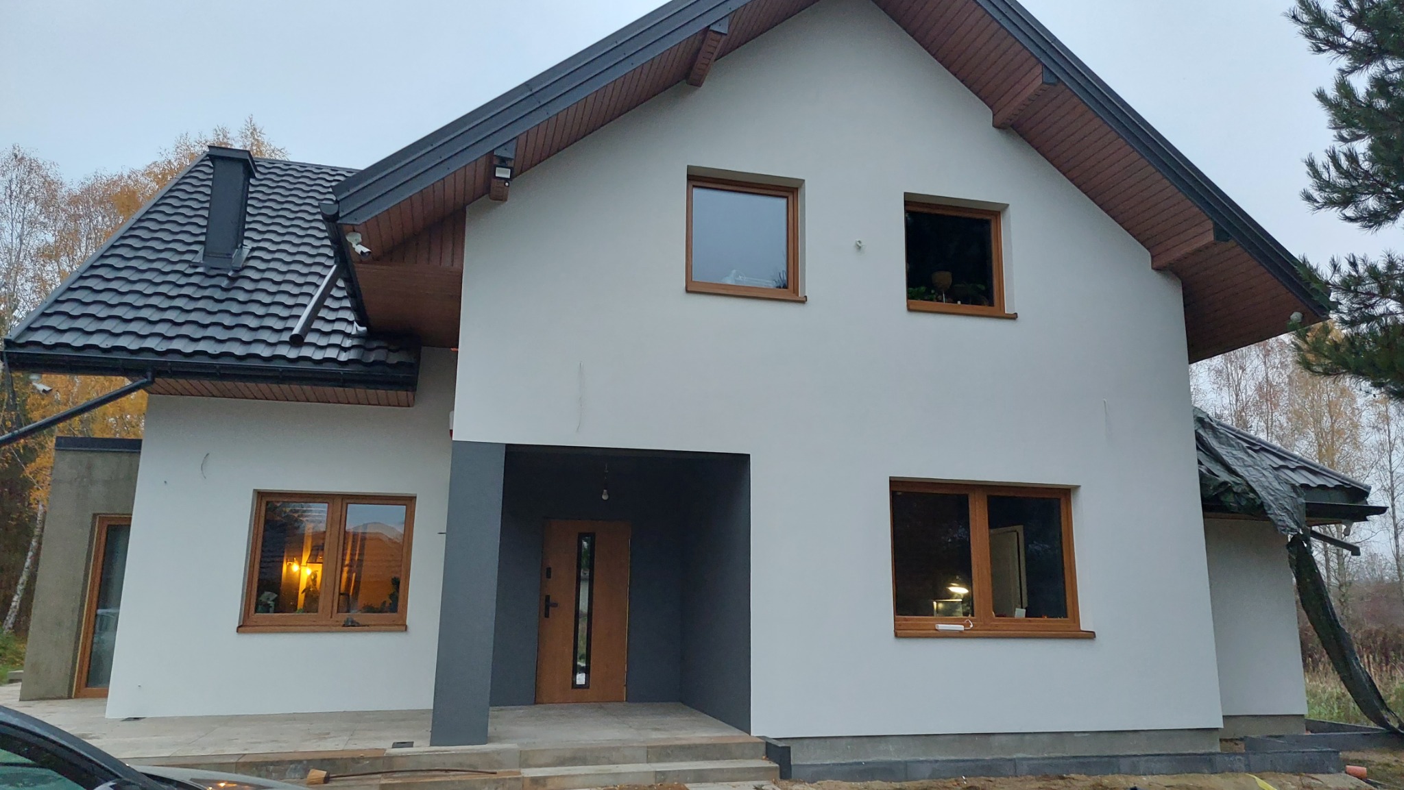 Dwukondygnacyjny dom jednorodzinny z szarą elewacją, brązowymi oknami i dachem z czarnej blachodachówki, widok z zewnątrz w pochmurny dzień.