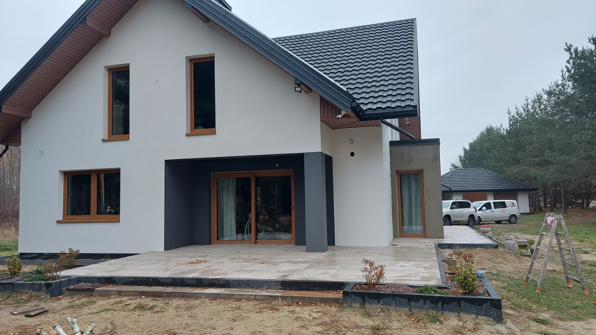 Nowoczesny dom jednorodzinny z białą elewacją, grafitowym dachem i drewnianymi oknami, widoczny fragment tarasu z płyt kamiennych i niedokończone prace wykończeniowe.