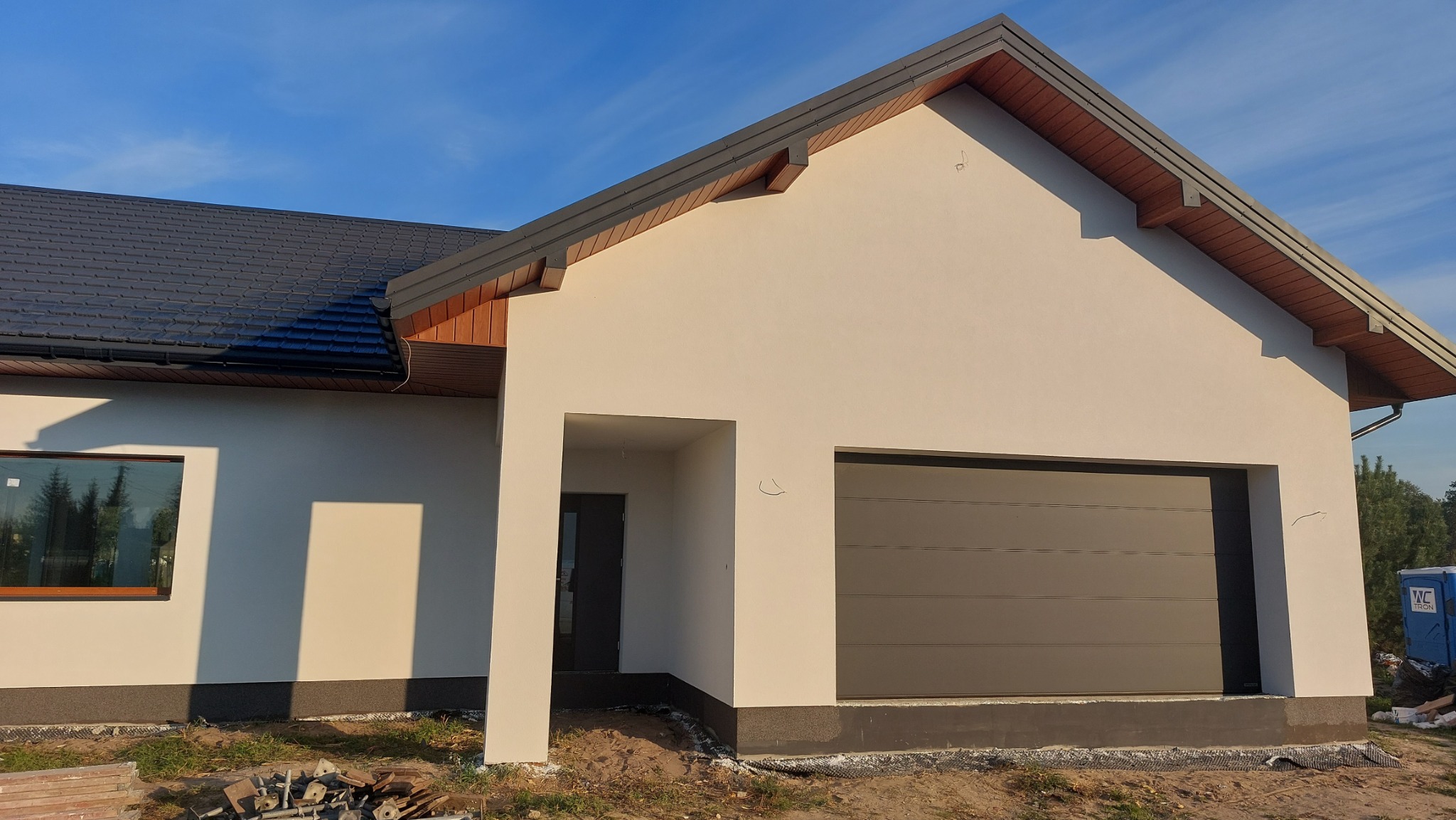 Nowoczesny dom jednorodzinny z jasną elewacją, grafitowym dachem i bramą garażową, widok z zewnątrz z elementami placu budowy.