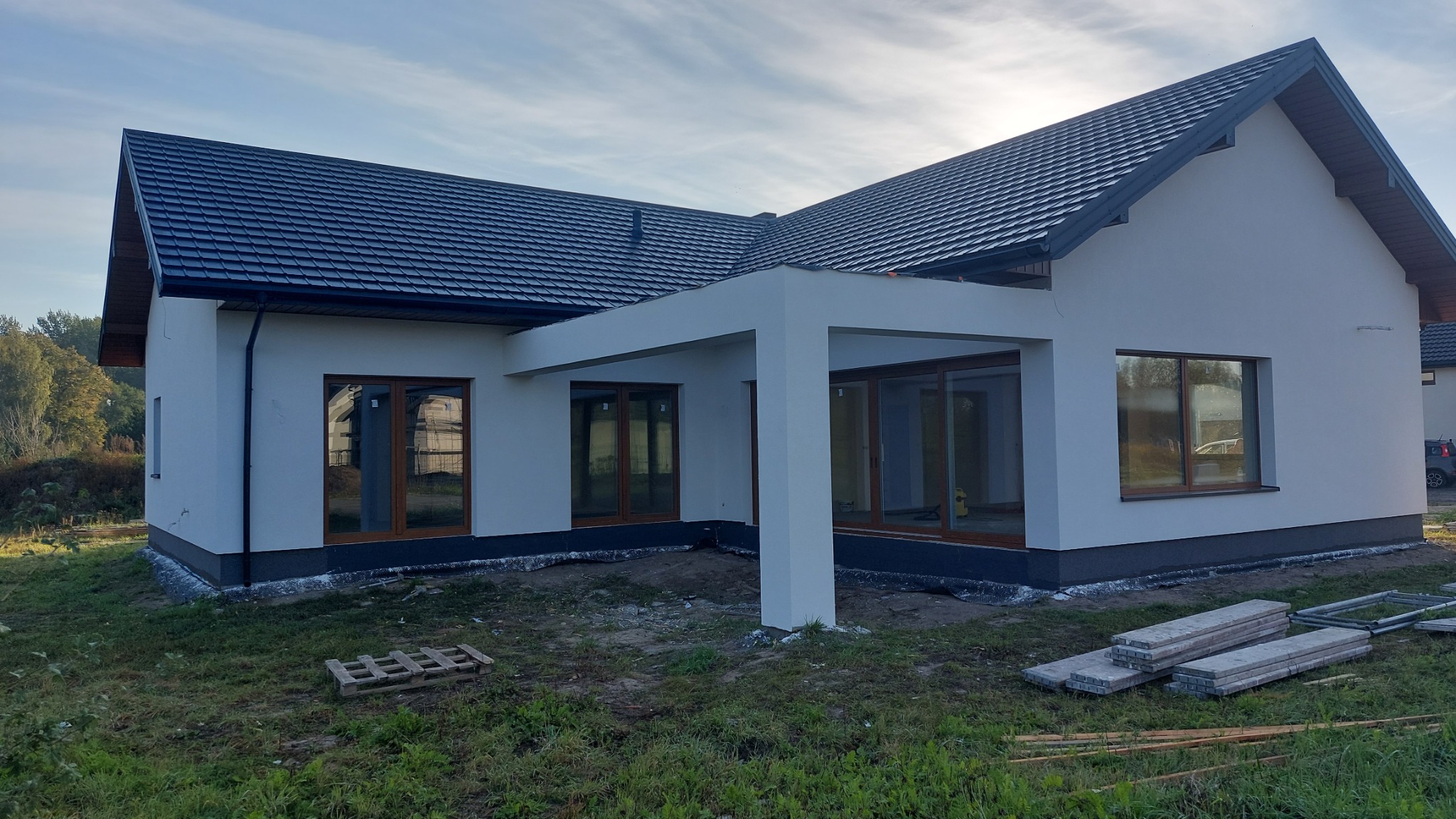 Nowoczesny dom jednorodzinny z białą elewacją, grafitowym dachem i jasną, otwartą konstrukcją tarasu, widoczne drewniane okna i paleta z materiałami na trawniku.