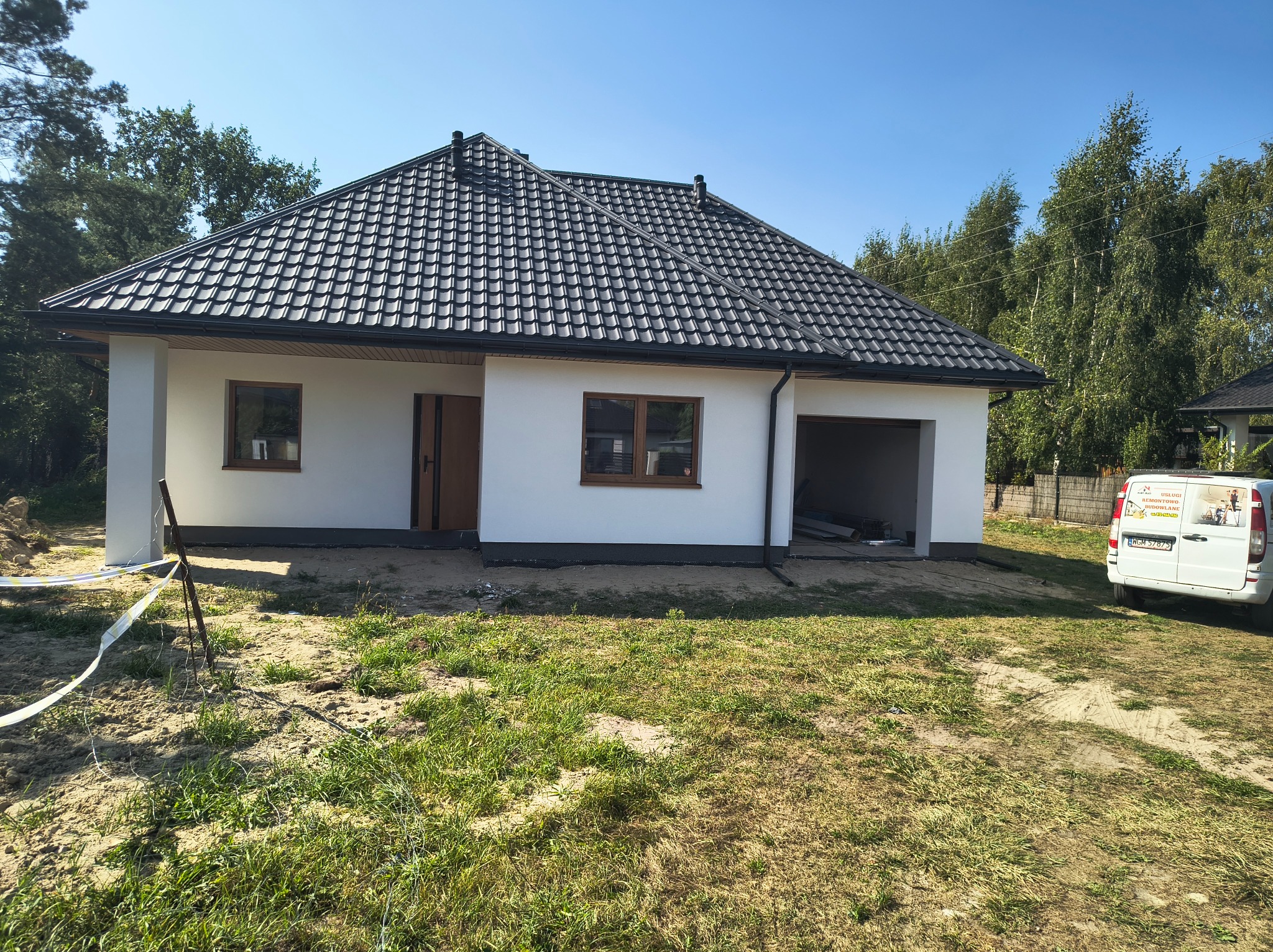 Biały dom jednorodzinny z ciemną dachówką, widok z zewnątrz. Częściowo wykończona elewacja, teren wokół domu w trakcie zagospodarowania. Na podjeździe stoi biały bus.