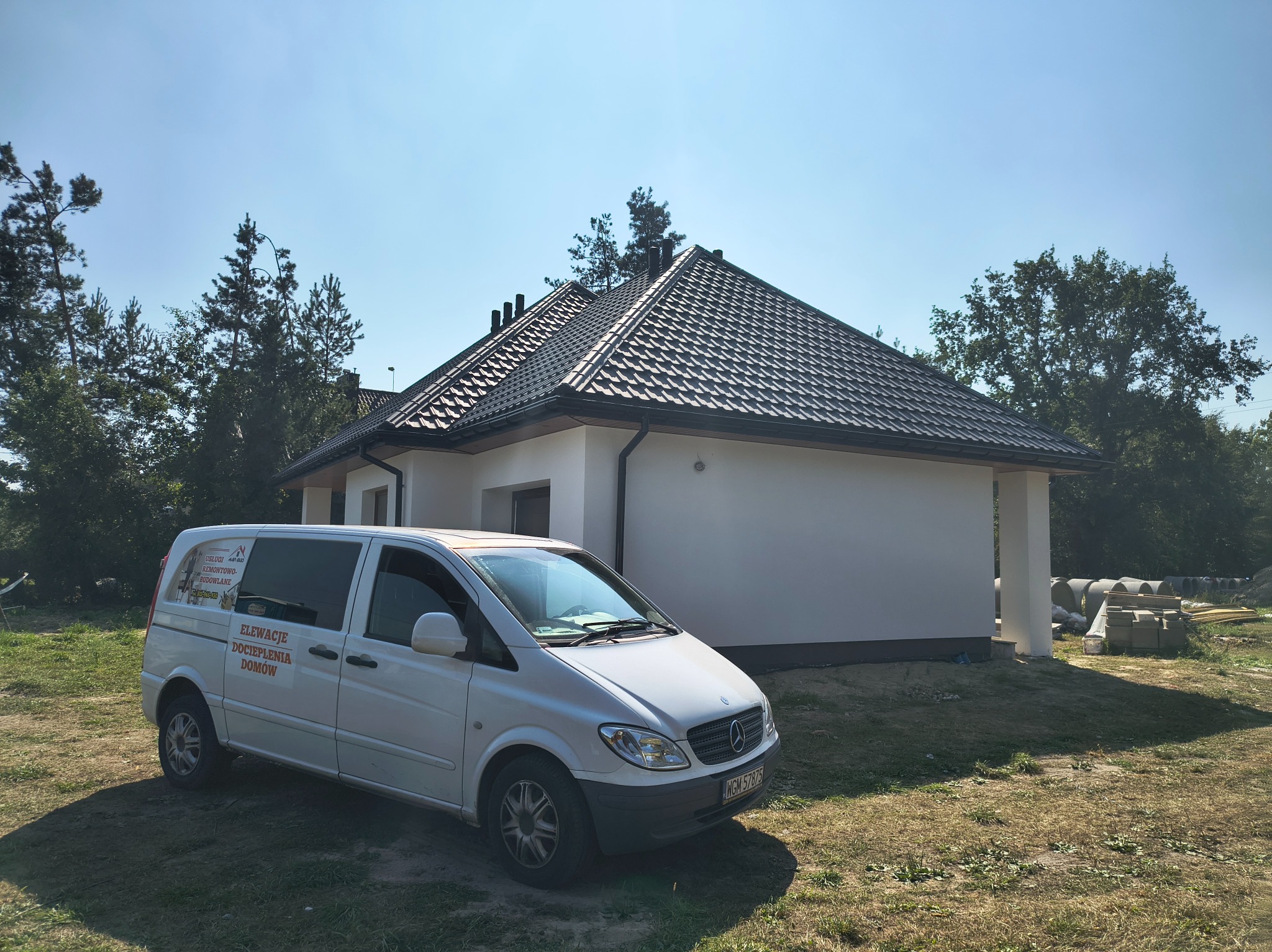 Biały, otynkowany dom jednorodzinny z ciemną dachówką, na pierwszym planie firmowy bus z napisem 'Elewacje Docieplenia Domów'. Widoczne materiały budowlane obok domu.