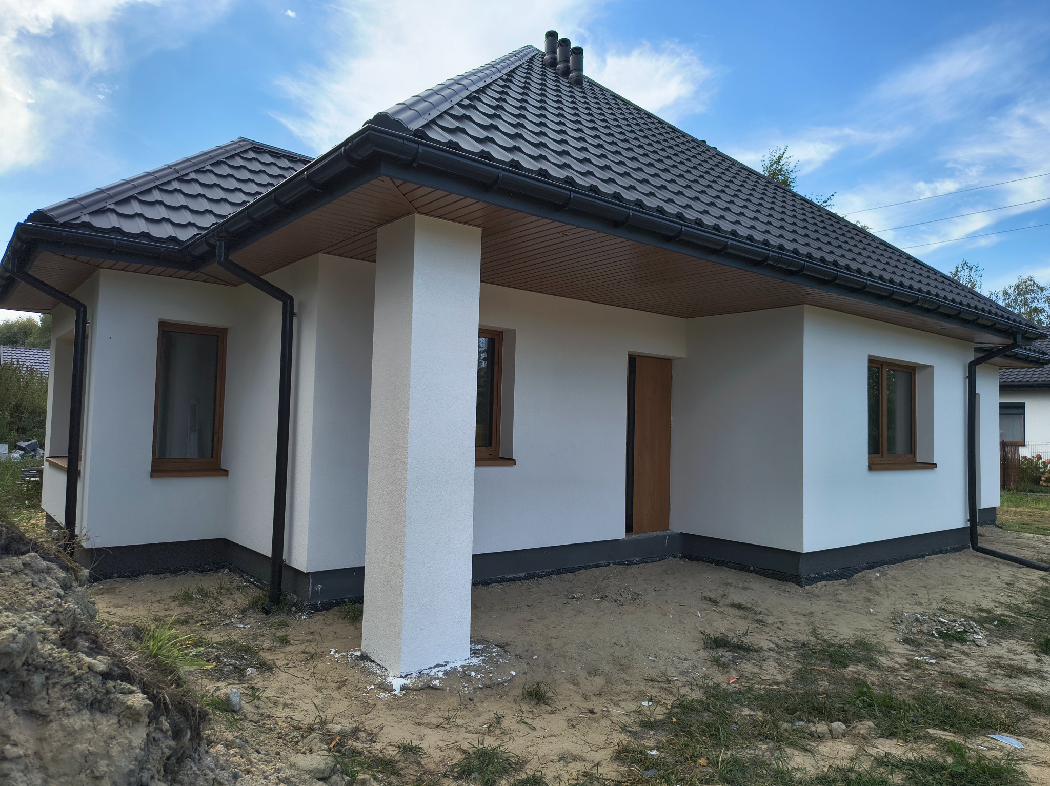 Biały dom jednorodzinny z ciemną dachówką i drewnianymi oknami w słoneczny dzień. Widoczny fragment elewacji i otoczenie w stanie surowym.
