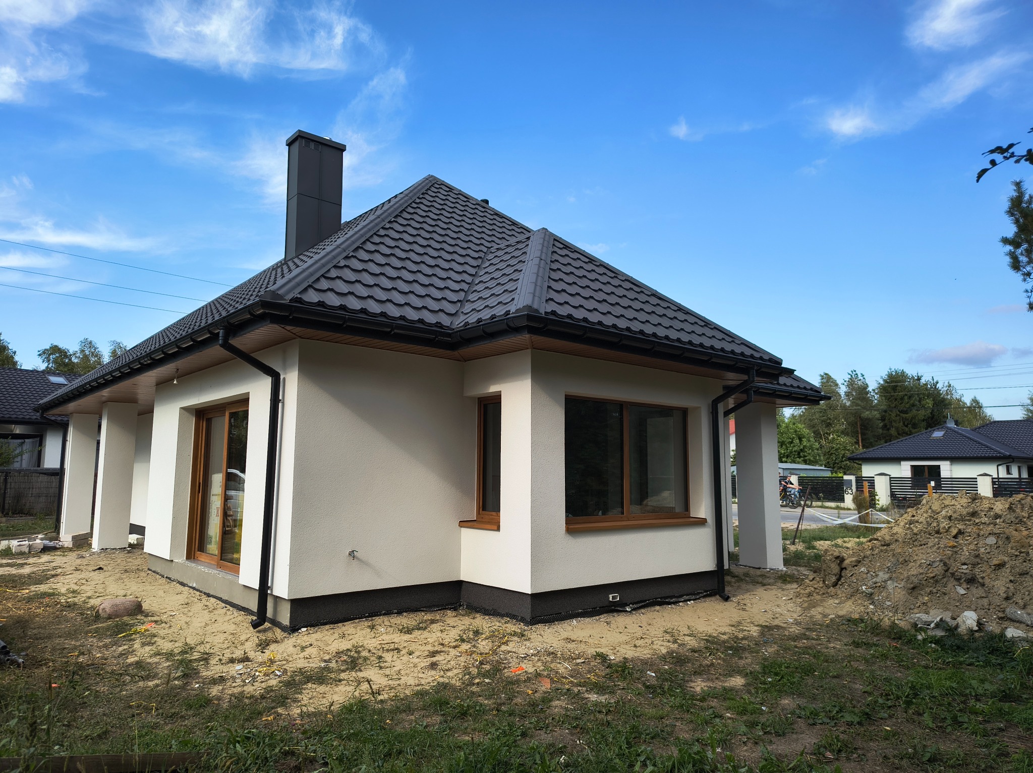 Nowoczesny dom jednorodzinny z jasną elewacją i ciemną dachówką na tle błękitnego nieba. Widoczne detale wykończenia i otoczenie domu.