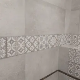 Wykończona ściana łazienki z jasnymi, matowymi płytkami imitującymi kamień oraz dekoracyjnym pasem płytek z geometrycznymi wzorami w odcieniach beżu i szarości, wykończona ciemnobrązową listwą...