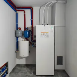 Biała pompa ciepła Atlantic z podłączonym zasobnikiem wody Gelmet i naczyniami wyrównawczymi w kotłowni z instalacją grzewczą z rur miedzianych i plastikowych.