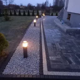 Oświetlona ścieżka z kostki brukowej i kamienia prowadząca wzdłuż domu z widocznym ogrodem w tle, oświetlona lampami ogrodowymi w kształcie słupków.