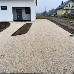 Nowo wysypany żwirowy podjazd do domu z obrzeżami, z przygotowanymi rabatami na zieleń, widok na sąsiedni dom i błotniste podłoże.