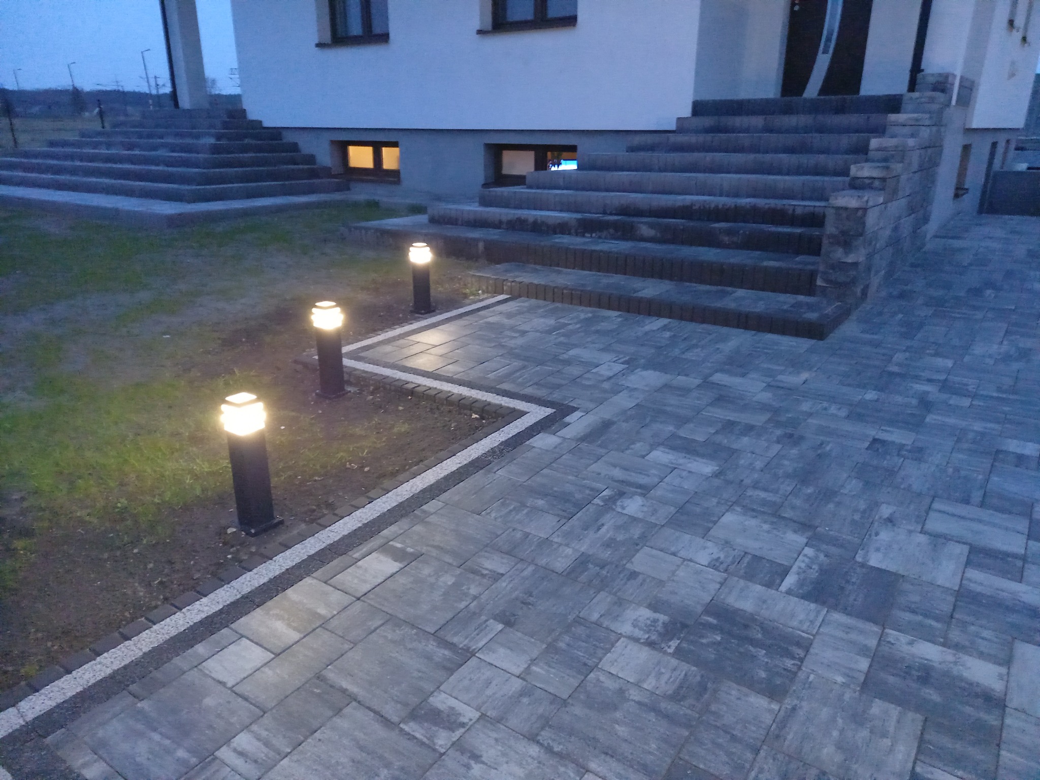 Nowoczesna posesja z szarej kostki brukowej, oświetlona trzema lampami stojącymi, z widocznymi schodami wejściowymi do domu.