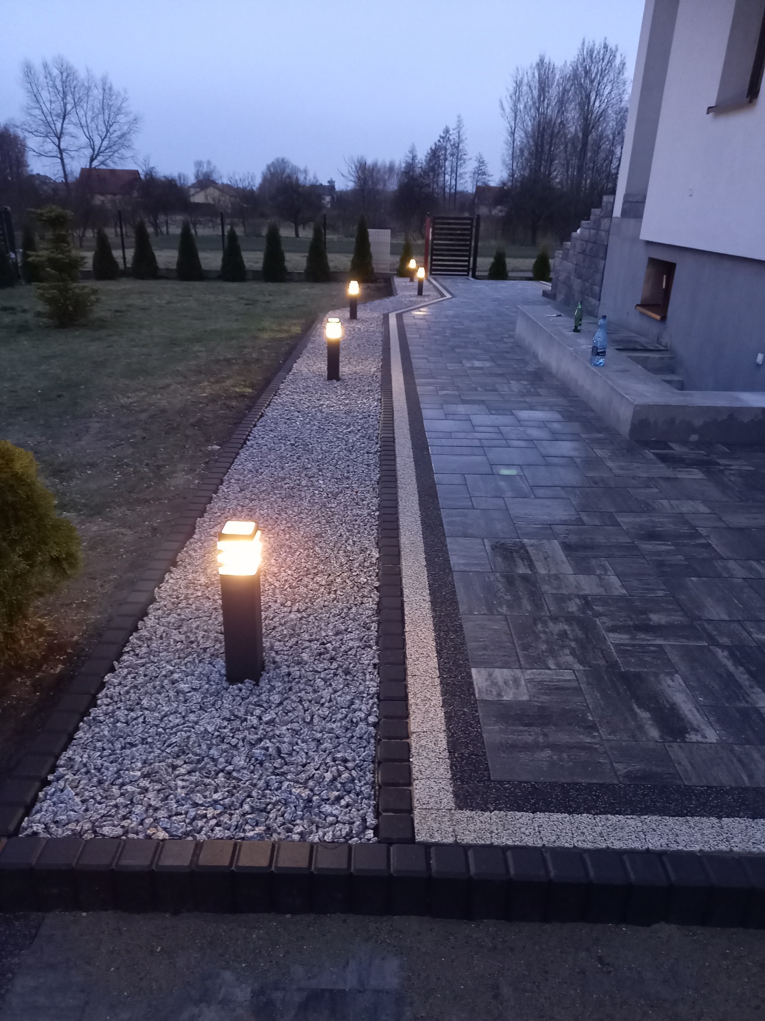 Oświetlona ścieżka z kostki brukowej i kamienia prowadząca wzdłuż domu z widocznym ogrodem w tle, oświetlona lampami ogrodowymi w kształcie słupków.