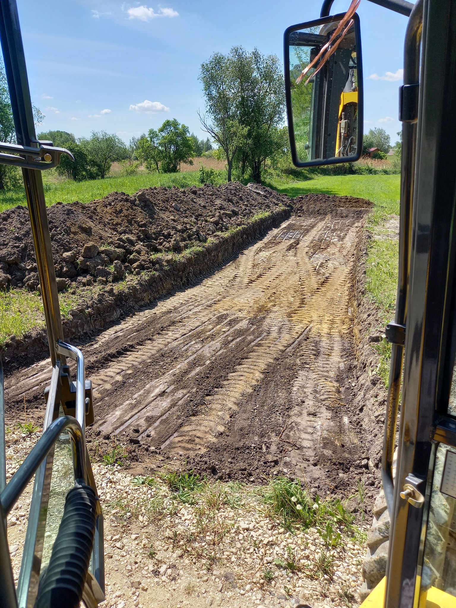 Widok z kabiny koparki na wykopany rów w ziemi, widoczne ślady opon, hałda ziemi po lewej stronie, lustro wsteczne z odbiciem maszyny, na horyzoncie zieleń drzew i błękitne niebo.