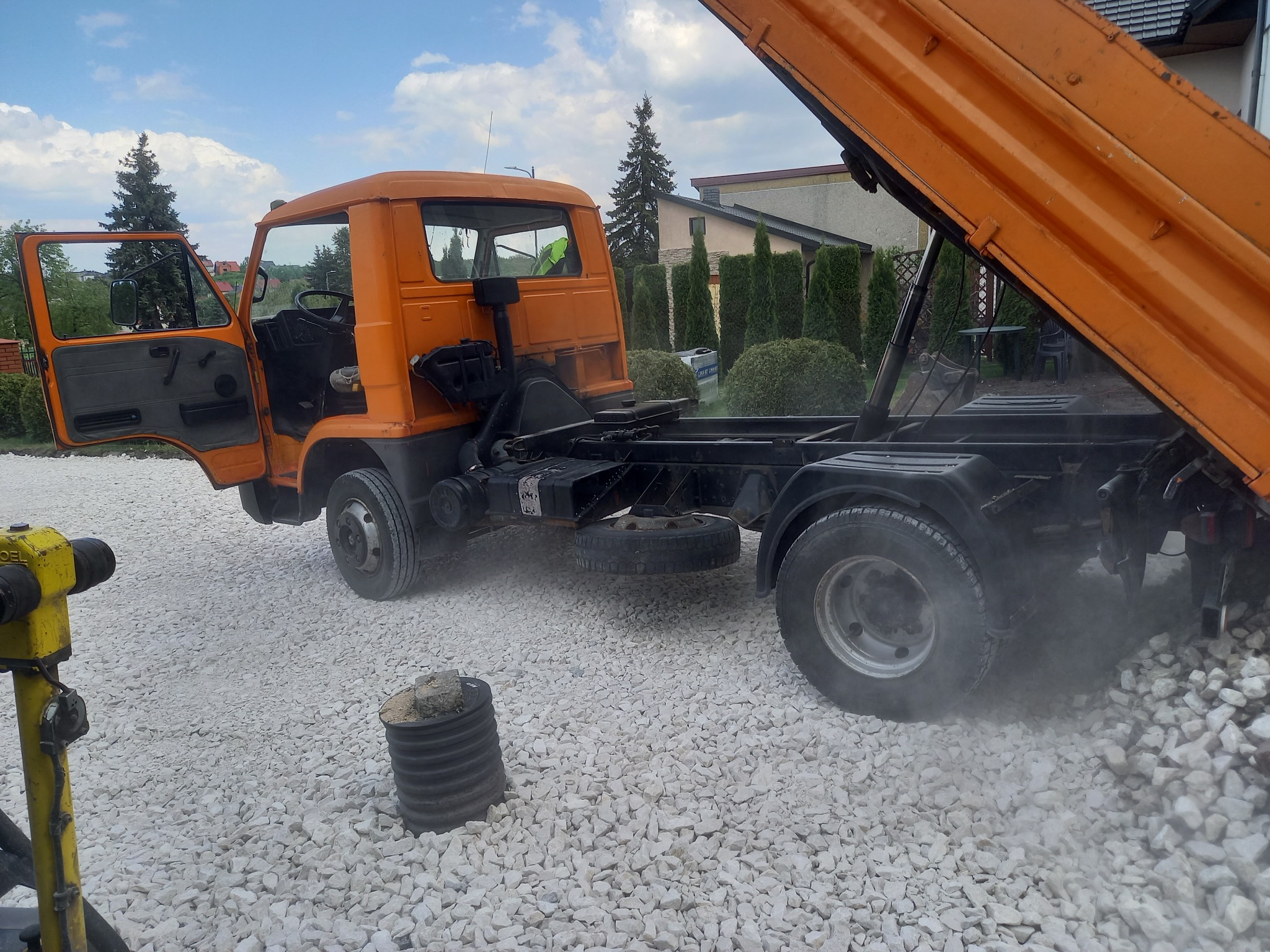 Pomarańczowa wywrotka z otwartą kabiną i podniesioną skrzynią, wysypująca biały tłuczeń na utwardzany teren. W tle widoczny dom i zielona roślinność.