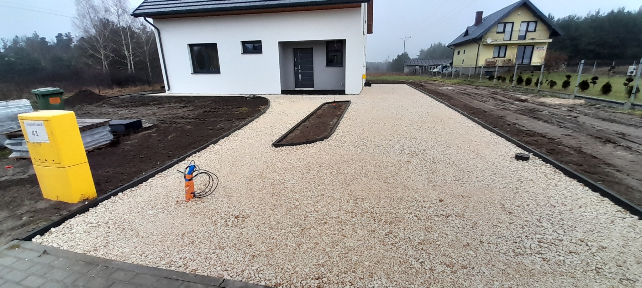 Podjazd wysypany jasnym żwirem, obrzeża z tworzywa, widoczny fragment trawnika, w tle dom z białym tynkiem i szarą stolarką okienną i drzwiową, oraz drugi dom z żółtą elewacją.