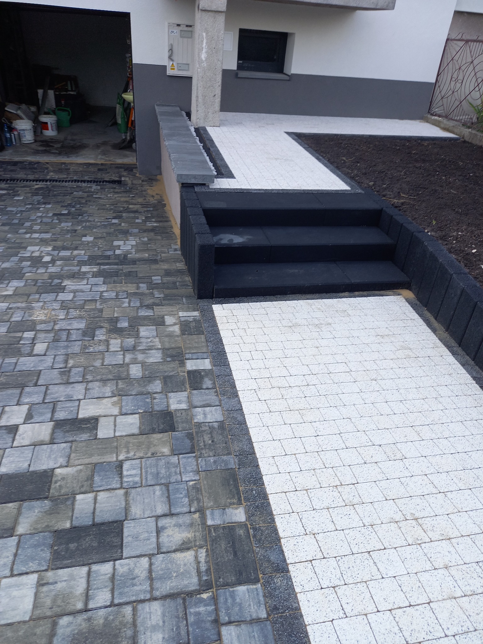 Ułożona z kostki brukowej ścieżka w dwóch odcieniach szarości, prowadząca od garażu do schodów wejściowych, z widoczną kratką odpływową i świeżo obsianą ziemią po bokach.