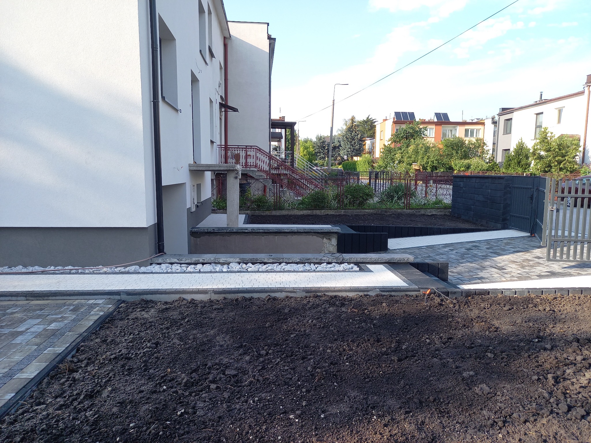 Nowo ułożona kostka brukowa w odcieniach szarości i bieli, obok przygotowany teren pod nasadzenia, widoczny fragment budynku mieszkalnego z czerwonymi schodami i metalowym ogrodzeniem w tle.