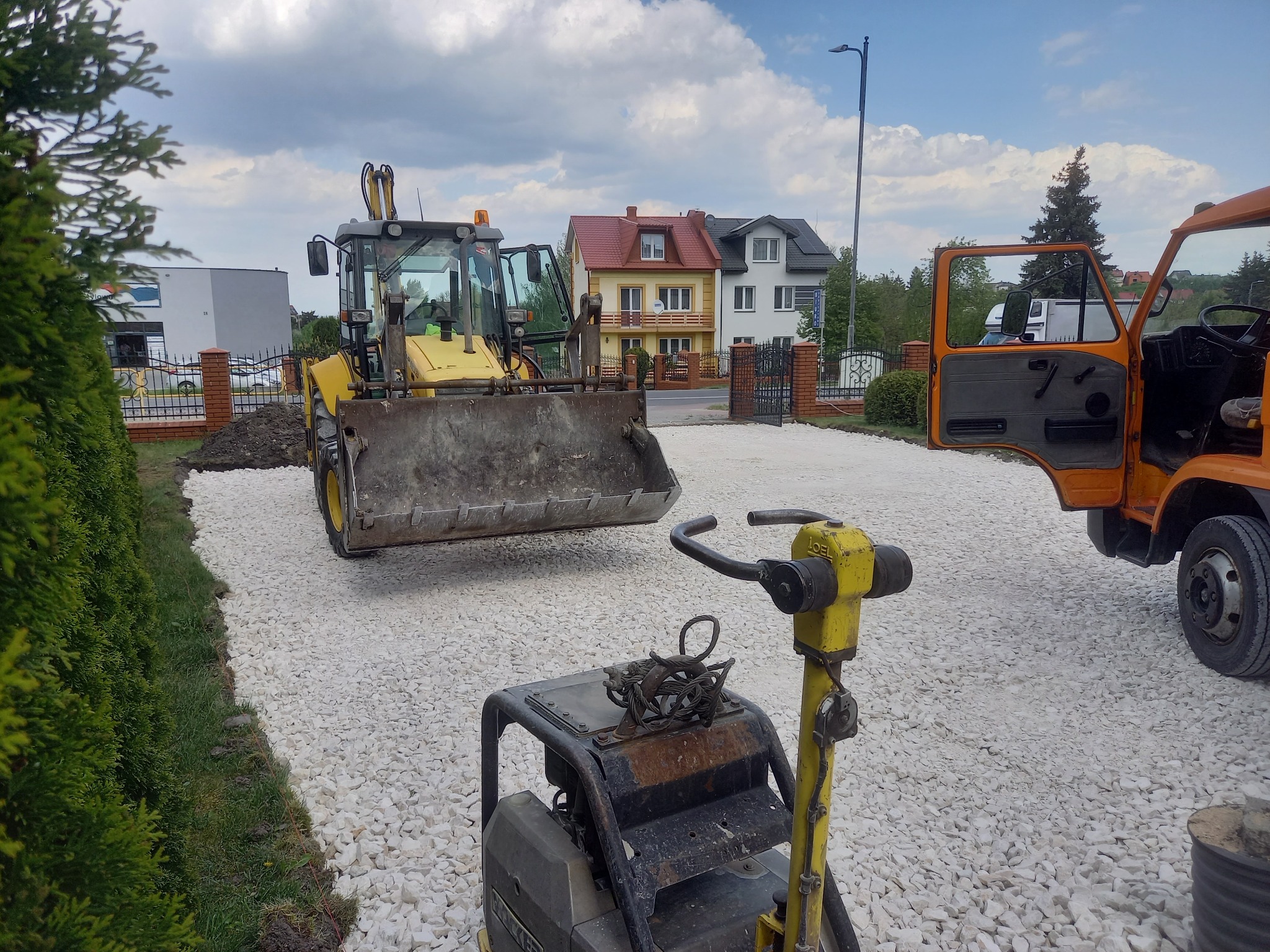 Żółta koparko-ładowarka z otwartą kabiną, ubijarka wibracyjna LOEL i pomarańczowa ciężarówka na podłożu z białego kruszywa na tle domów jednorodzinnych.