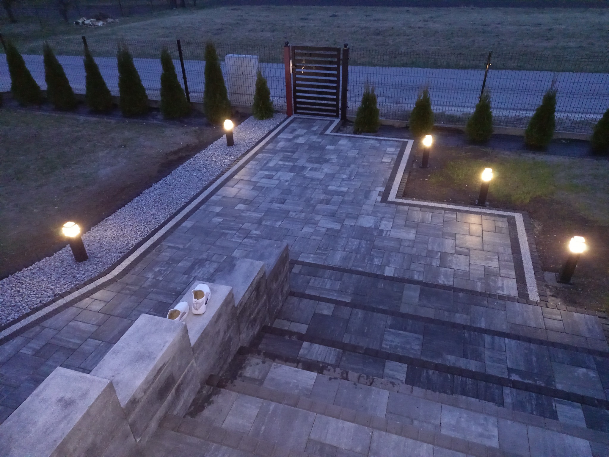 Oświetlona, nowa ścieżka z kostki brukowej prowadząca do furtki, z parą białych butów leżących na schodach wejściowych, otoczona ozdobnymi krzewami i kamieniami.