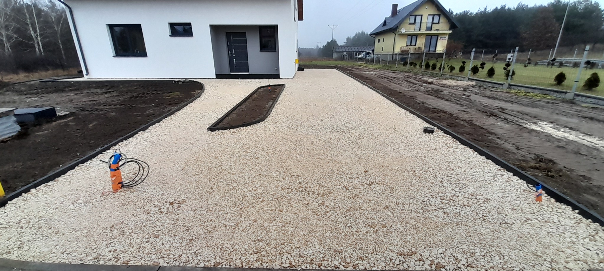 Nowo wysypany żwirowy podjazd do domu z obrzeżami, z przygotowanymi rabatami na zieleń, widok na sąsiedni dom i błotniste podłoże.