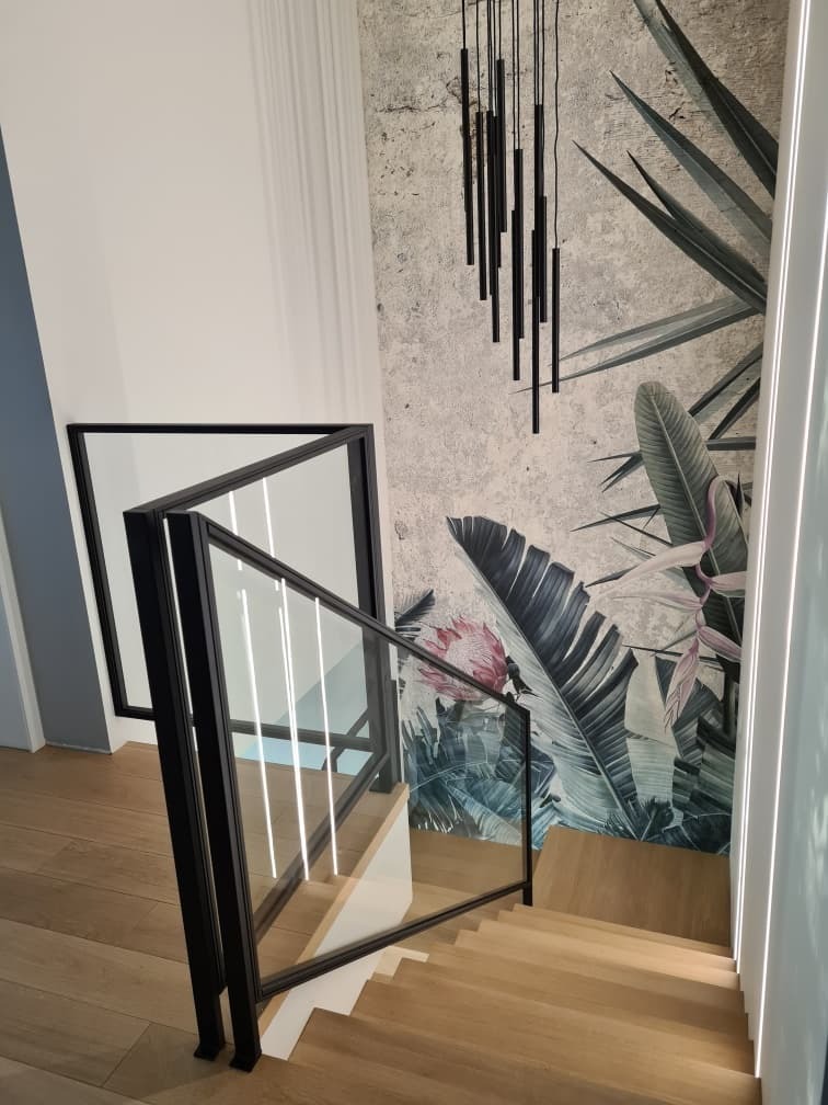 Schody z drewnianymi stopniami i szklaną balustradą z czarnymi ramami, oświetlenie LED wzdłuż ściany, na ścianie fototapeta z motywem tropikalnych liści i wisząca lampa z czarnych rurek.