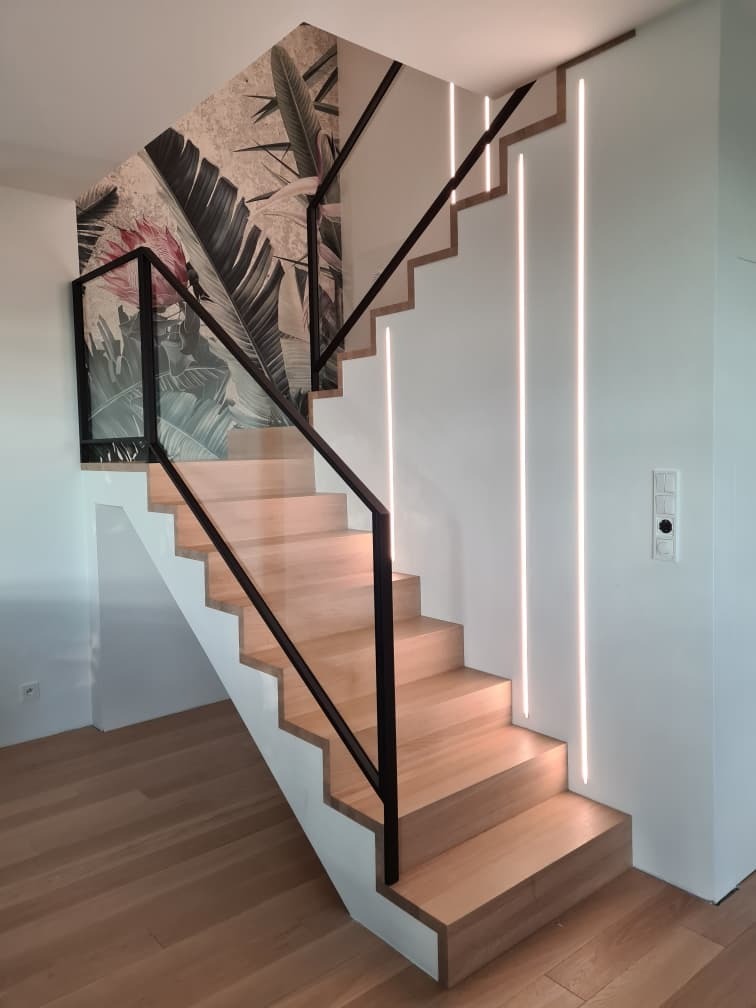 Drewniane schody z czarną balustradą i oświetleniem LED w ścianie, ozdobna tapeta z motywem roślinnym na ścianie.