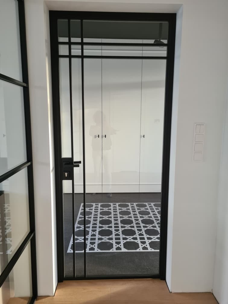Czarne drzwi z szybami w stylu loftowym, widok na białą szafę i czarno-białą mozaikę podłogową w przedpokoju.