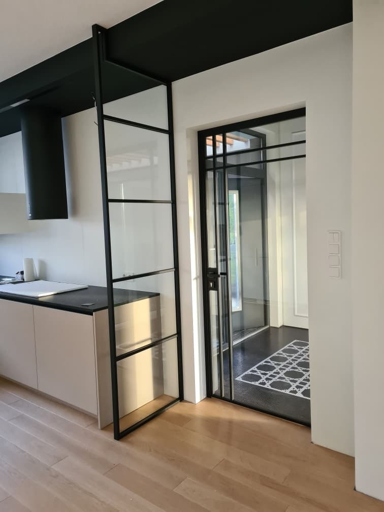 Nowoczesne wnętrze z przeszklonymi drzwiami przesuwnymi z czarnymi szprosami, oddzielającymi kuchnię z białymi szafkami i czarnym okapem od przedpokoju z wzorzystym dywanem.