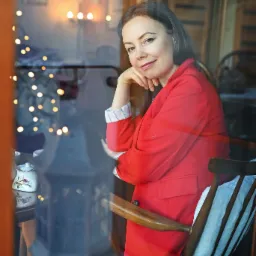 Uśmiechnięta kobieta w czerwonej marynarce siedzi przy stole z ceramicznym czajnikiem, widoczna przez szybę z refleksami światełek.