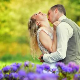 Uśmiechnięta panna młoda w koronkowej sukni, obejmowana przez pana młodego w szarej kamizelce na tle rozmytych fioletowych kwiatów i zieleni.