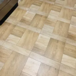 Beżowa podłoga z płytek imitujących drewno ułożonych w geometryczny wzór, widoczny fragment czarnej obudowy piekarnika.