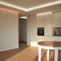 Jasne, przestronne wnętrze mieszkania z drewnianą podłogą, białymi ścianami i zabudową kuchenną z oświetleniem LED podsufitowym. Na pierwszym planie okrągły stół z drewnianym blatem i białymi...