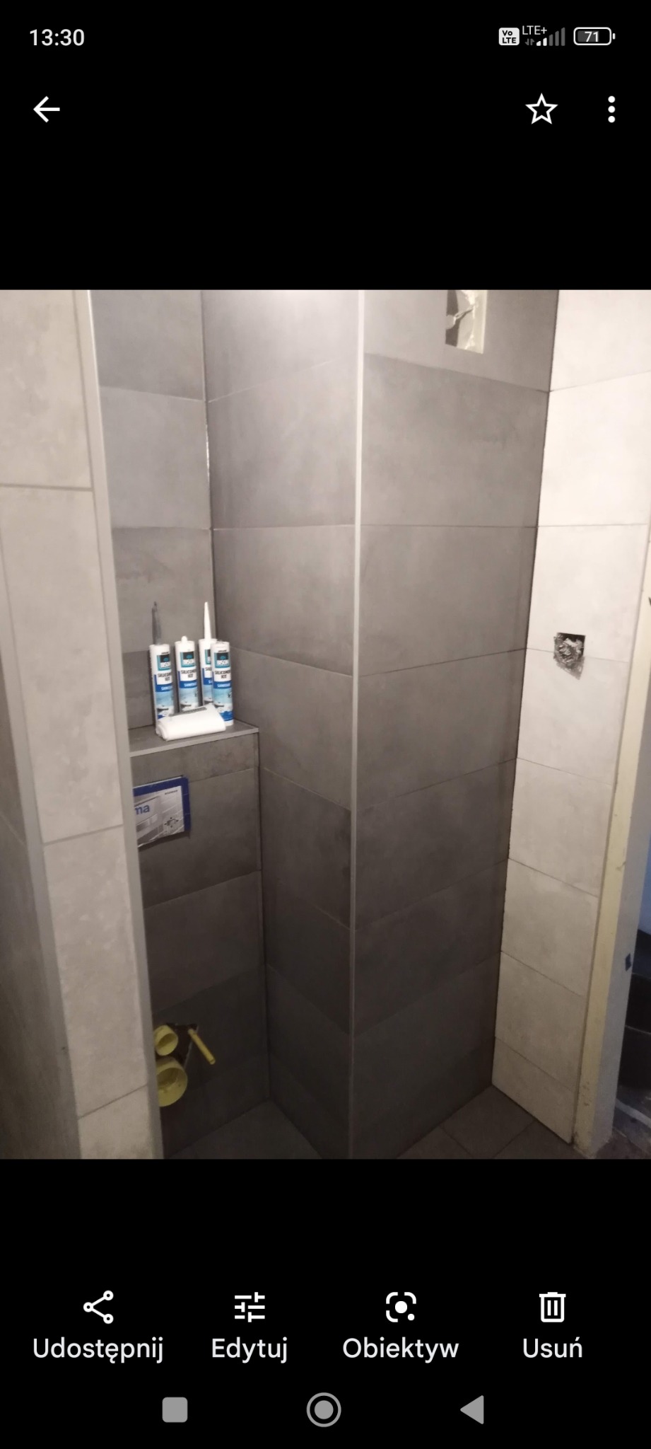 Wykończona wnęka prysznicowa z szarymi, poziomymi płytkami, biały silikon i biała ściereczka na półce, żółte akcesoria do montażu toalety w dolnej części wnęki.