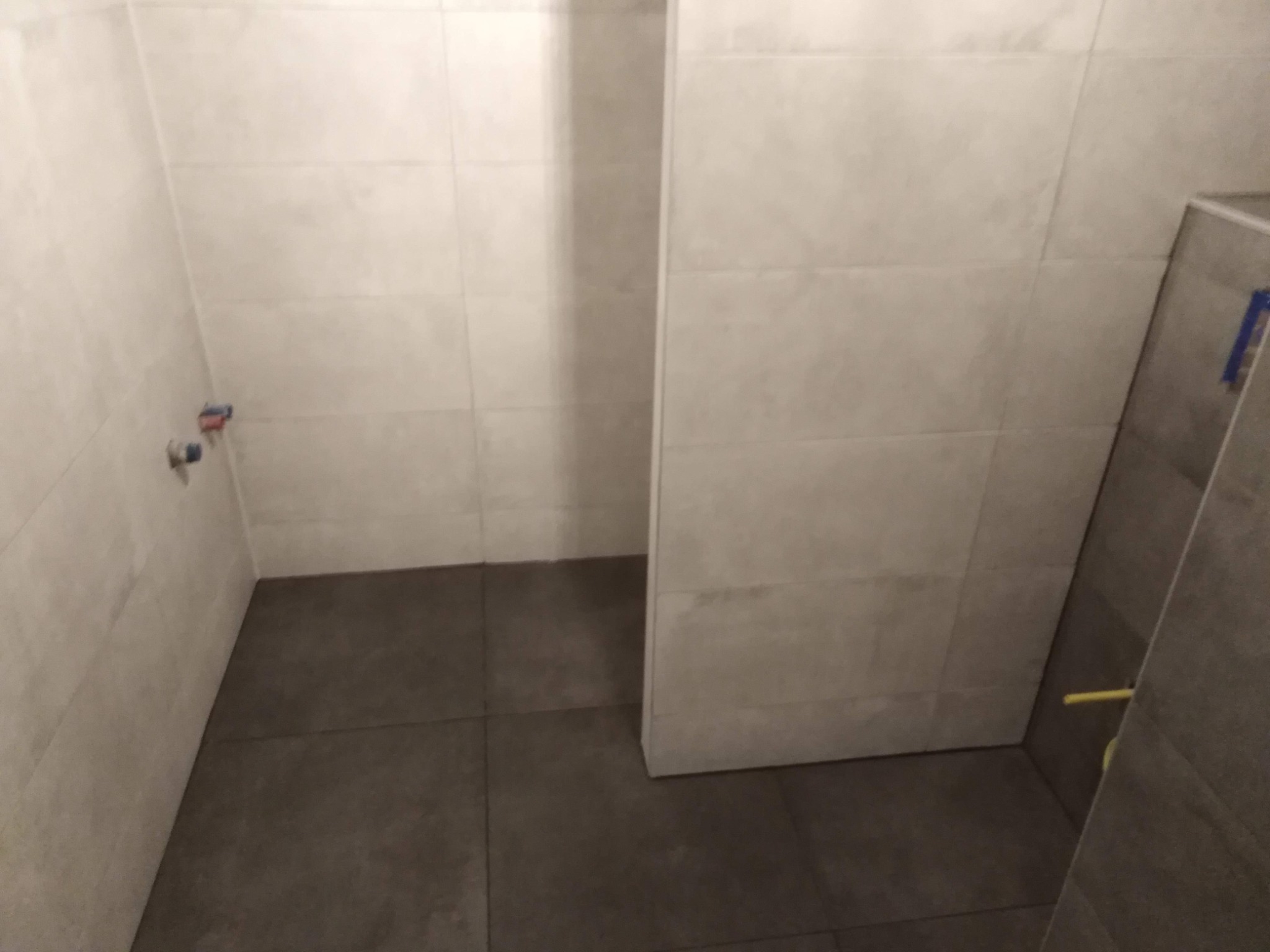 Wnętrze łazienki w trakcie remontu: jasne płytki ścienne, ciemne płytki podłogowe, widoczne przyłącza wodne, fragment kabiny prysznicowej zabezpieczonej taśmą.