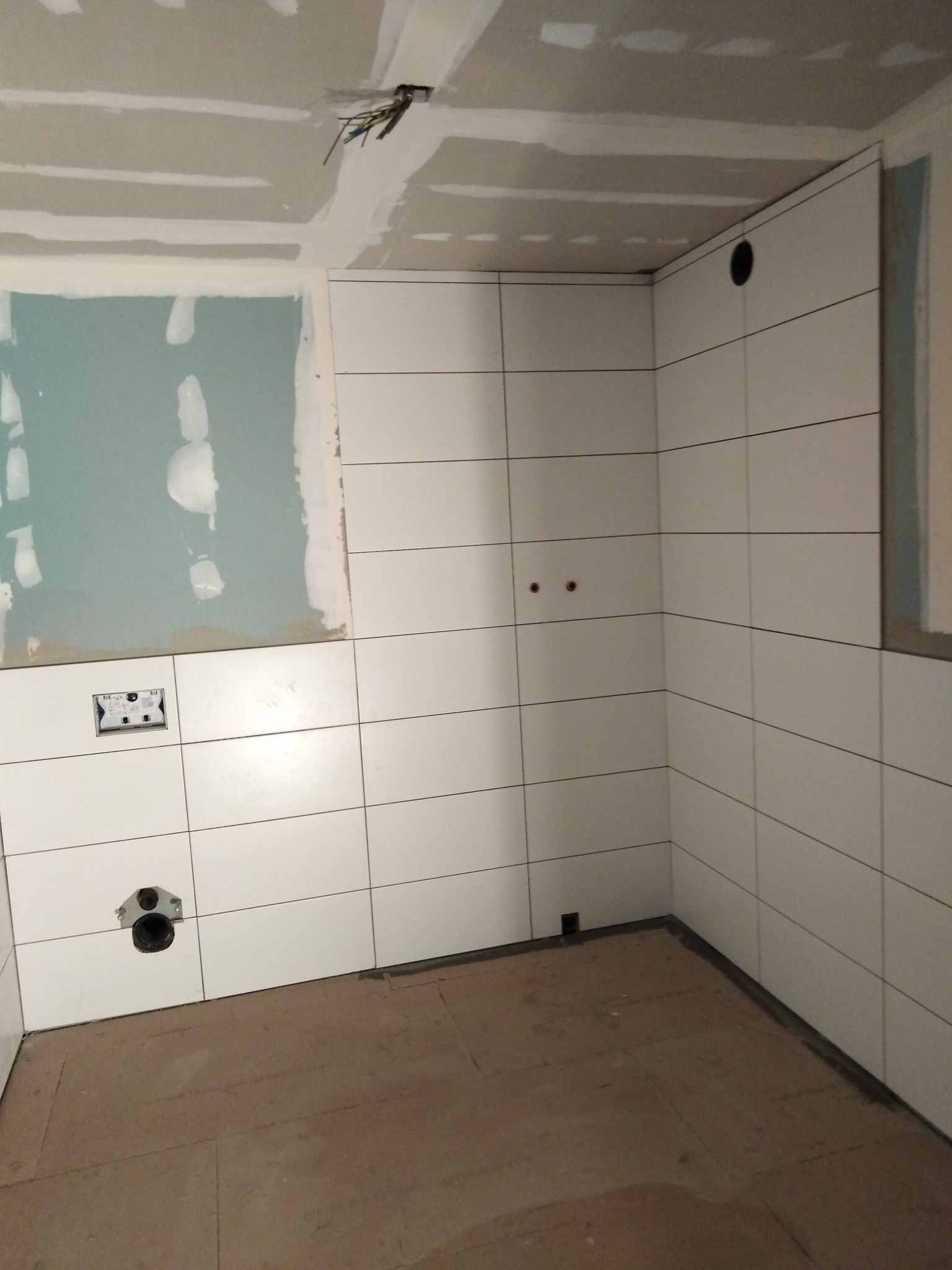 Wnętrze łazienki w trakcie wykańczania: białe, prostokątne płytki ułożone poziomo na ścianach, surowy sufit z płyt gipsowo-kartonowych, widoczne instalacje elektryczne i hydrauliczne, podłoga...