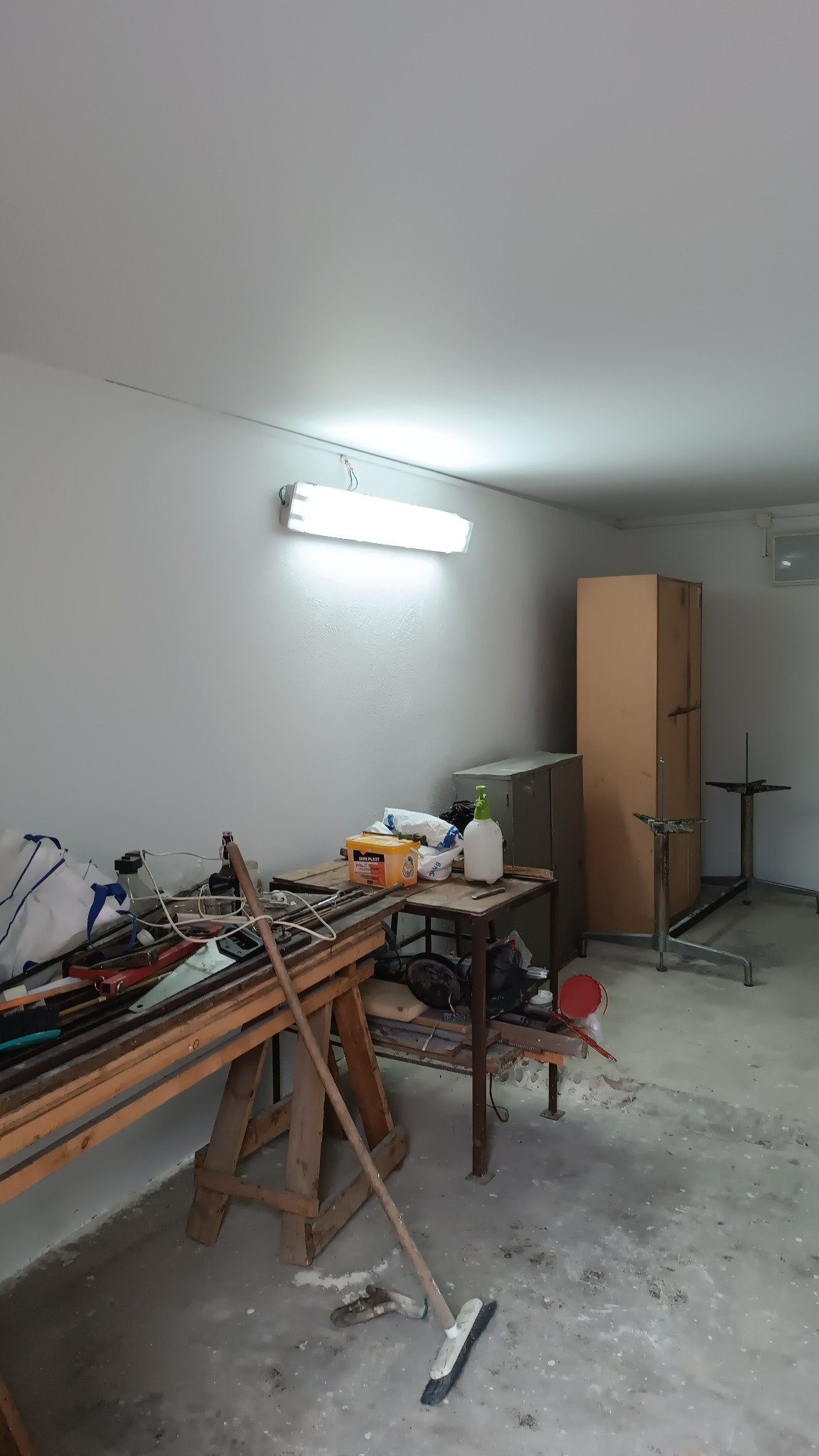Przygotowane pomieszczenie do prac wykończeniowych: widoczny stół roboczy z narzędziami, skrzynka, butelka z rozpylaczem, miotła oparta o stół, surowa podłoga i ściany, oświetlenie jarzeniowe.