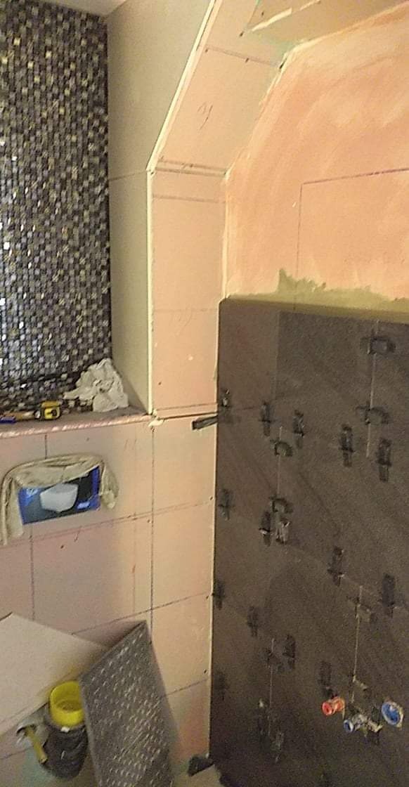 Układanie ciemnych płytek na ścianie w łazience, widoczne krzyżyki dystansowe, fragment mozaiki na sąsiedniej ścianie, narzędzia pracy na półce, prace w toku.