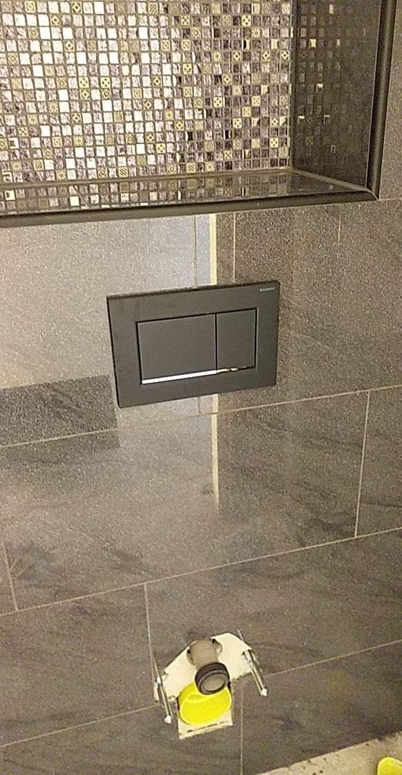 Szare, prostokątne płytki na ścianie łazienki, z czarnym panelem spłuczki i mozaiką w górnej części, widoczny montaż stelaża podtynkowego WC.