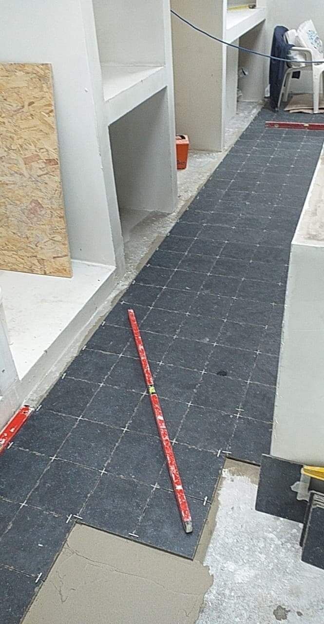 Układanie ciemnych płytek podłogowych w wąskim pomieszczeniu z wnękami, widoczna poziomica i krzyżyki dystansowe.