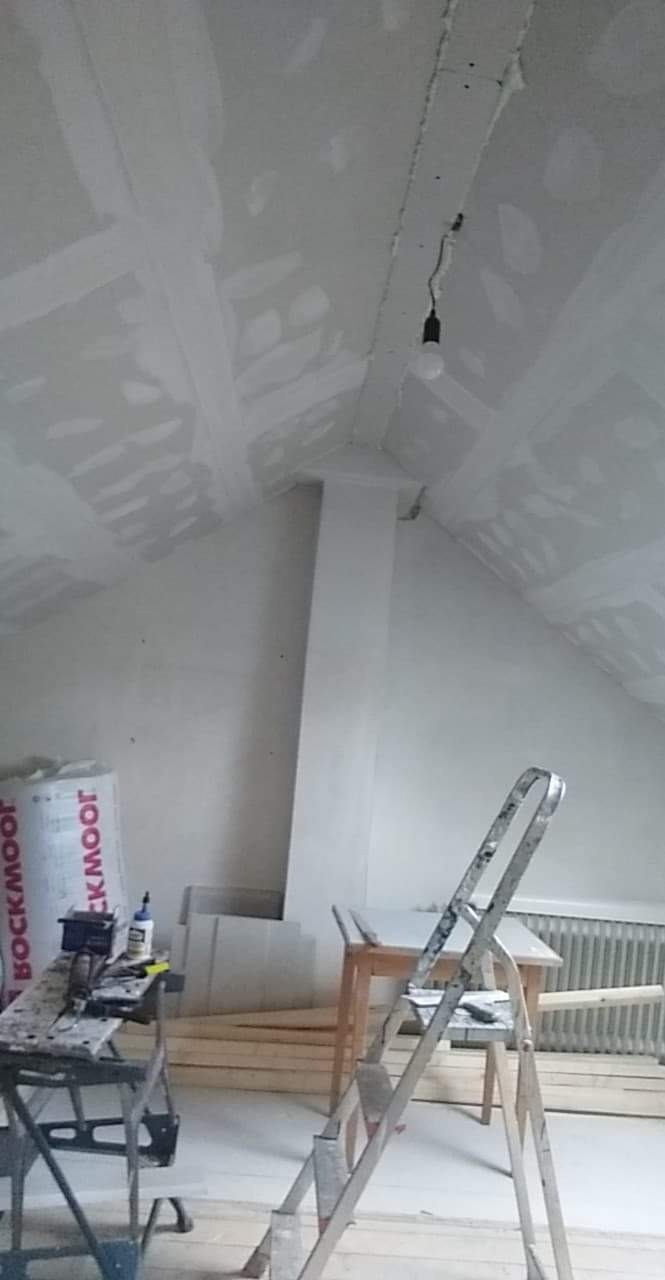 Poddasze w trakcie prac wykończeniowych: ściany i skosy sufitu pokryte gładzią, widoczne łączenia płyt gipsowych, metalowa drabina oparta o ścianę, podwieszona żarówka na kablu, materiały budowlane...