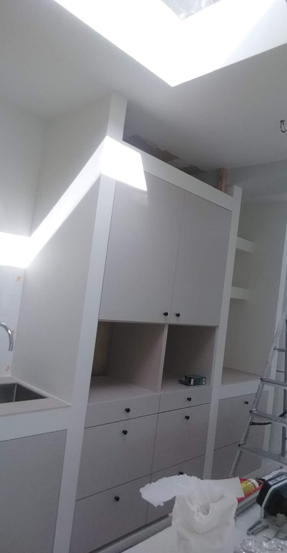 Biała zabudowa kuchenna pod skosem dachu z oknem, widoczny fragment zlewu, czarne okrągłe uchwyty, drabina oparta o ścianę, wnętrze w trakcie wykańczania.