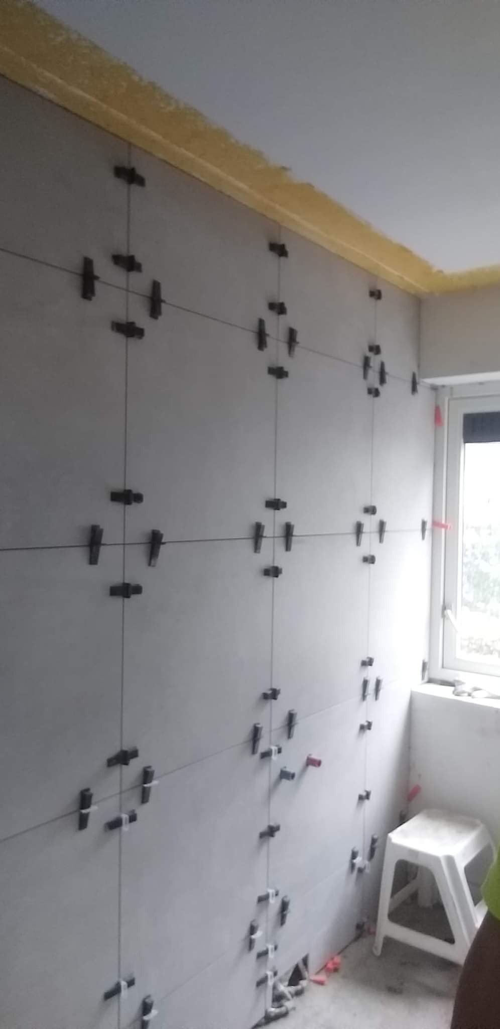 Świeżo ułożone, jasnoszare płytki ścienne z czarnymi krzyżykami dystansowymi i czerwonymi klinami, na górze żółta listwa maskująca, małe okno w rogu i biały stołek.