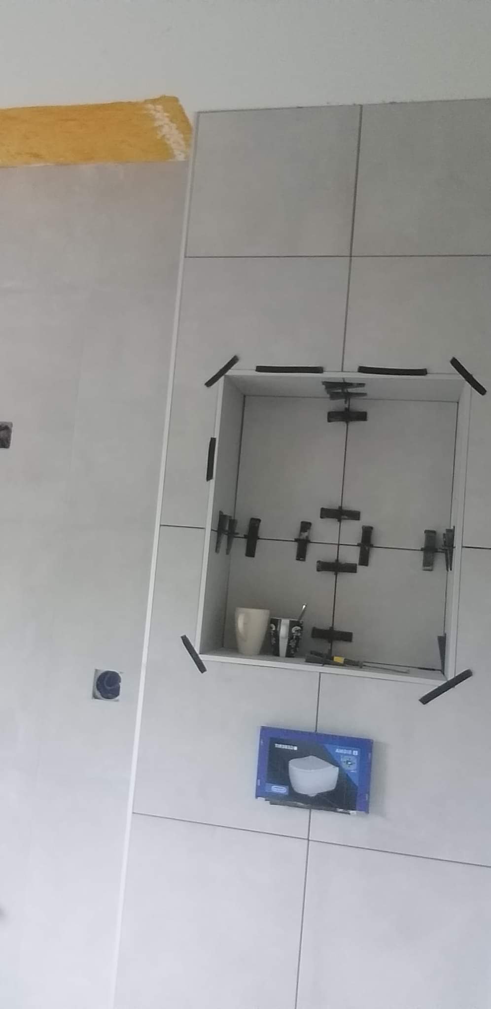 Świeżo ułożone, jasne płytki ceramiczne na ścianie z wnęką, zabezpieczone krzyżykami dystansowymi. W tle widoczne żółte zabezpieczenie na suficie i pudełko po ceramice. Wnęka zawiera kubek i przybory.