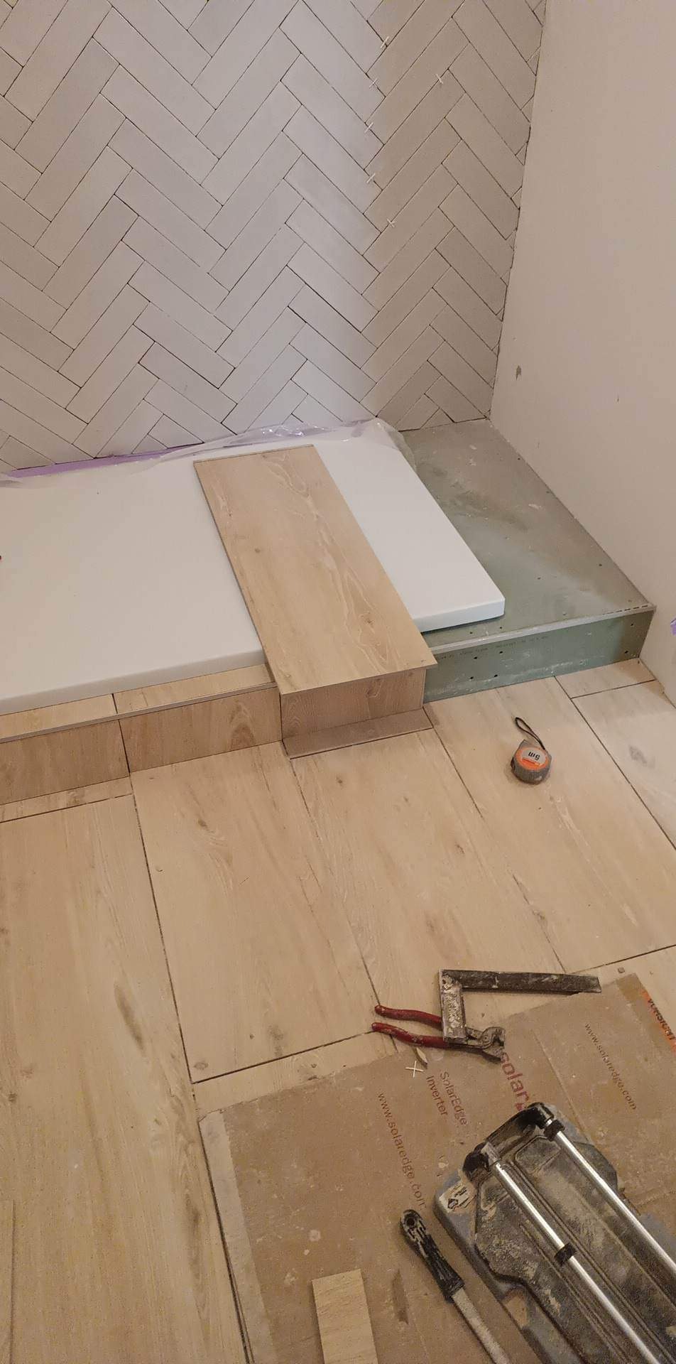 Łazienka w trakcie prac: Ściana wykończona białymi płytkami ułożonymi w jodełkę, podłoga z płytek imitujących drewno, fragment brodzika zabezpieczony folią, na podłodze narzędzia glazurnicze...