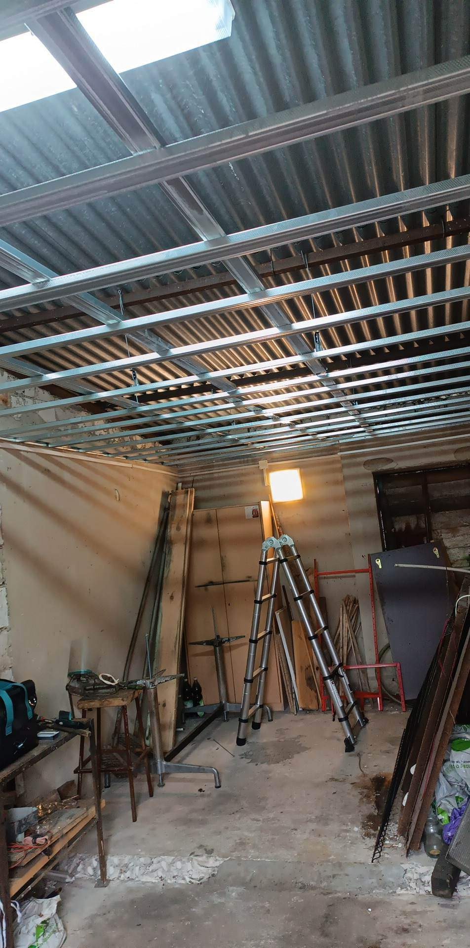 Metalowa konstrukcja sufitu podwieszanego z widoczną izolacją, oświetlona lampą warsztatową, z drabiny i narzędziami w tle.