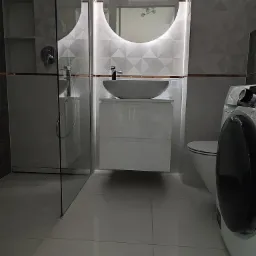 Nowoczesna łazienka z umywalką nablatową, okrągłym lustrem podświetlanym LED, kabiną prysznicową typu walk-in i pralką.