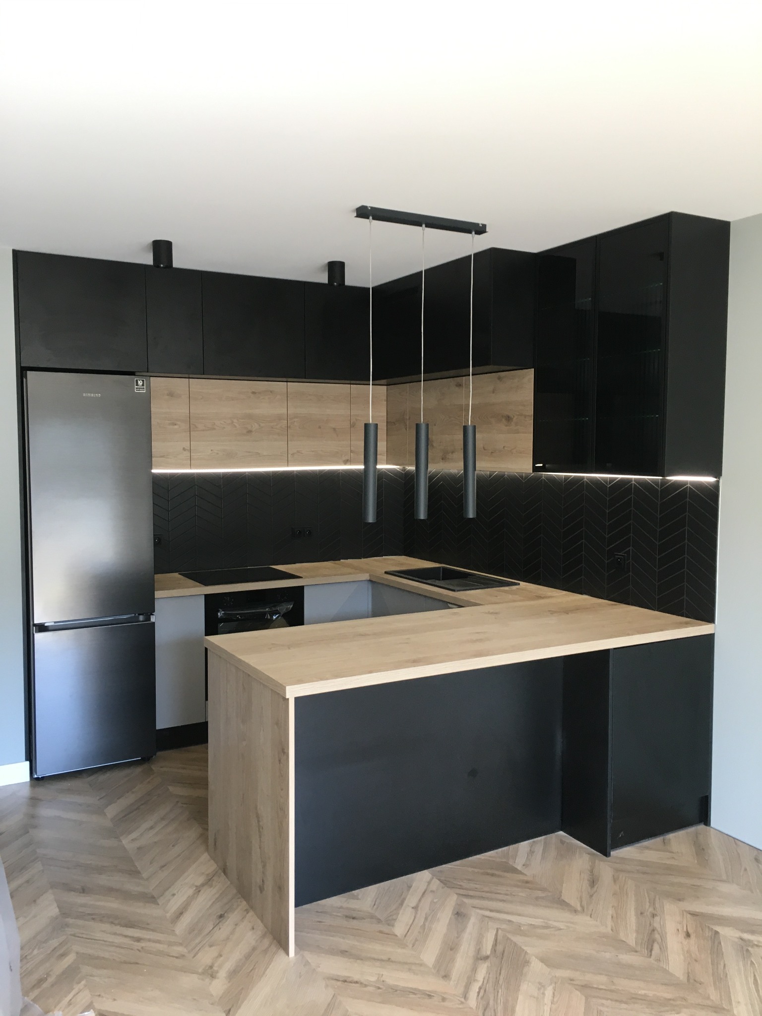 Nowoczesna kuchnia z czarnymi szafkami, drewnianymi frontami i blatem, oraz podłogą ułożoną w jodełkę, oświetlona wiszącymi lampami cylindrycznymi.