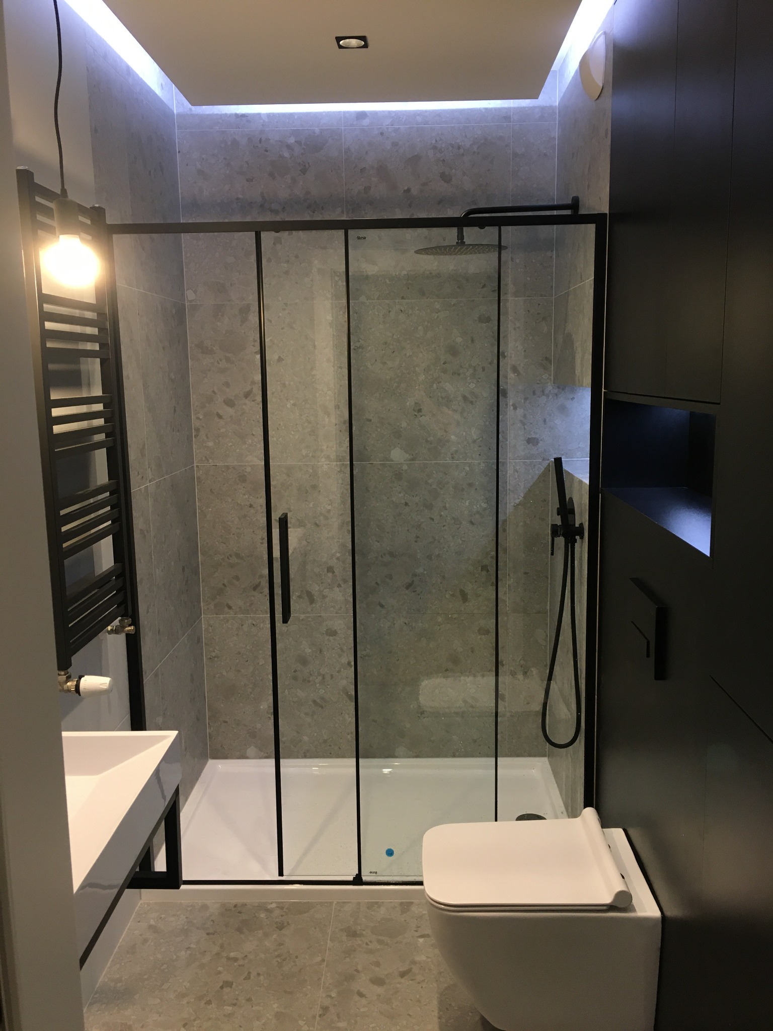 Nowoczesna łazienka z kabiną prysznicową typu walk-in z czarnymi profilami, podwieszaną toaletą, umywalką nablatową i grzejnikiem drabinkowym w ciemnej kolorystyce.