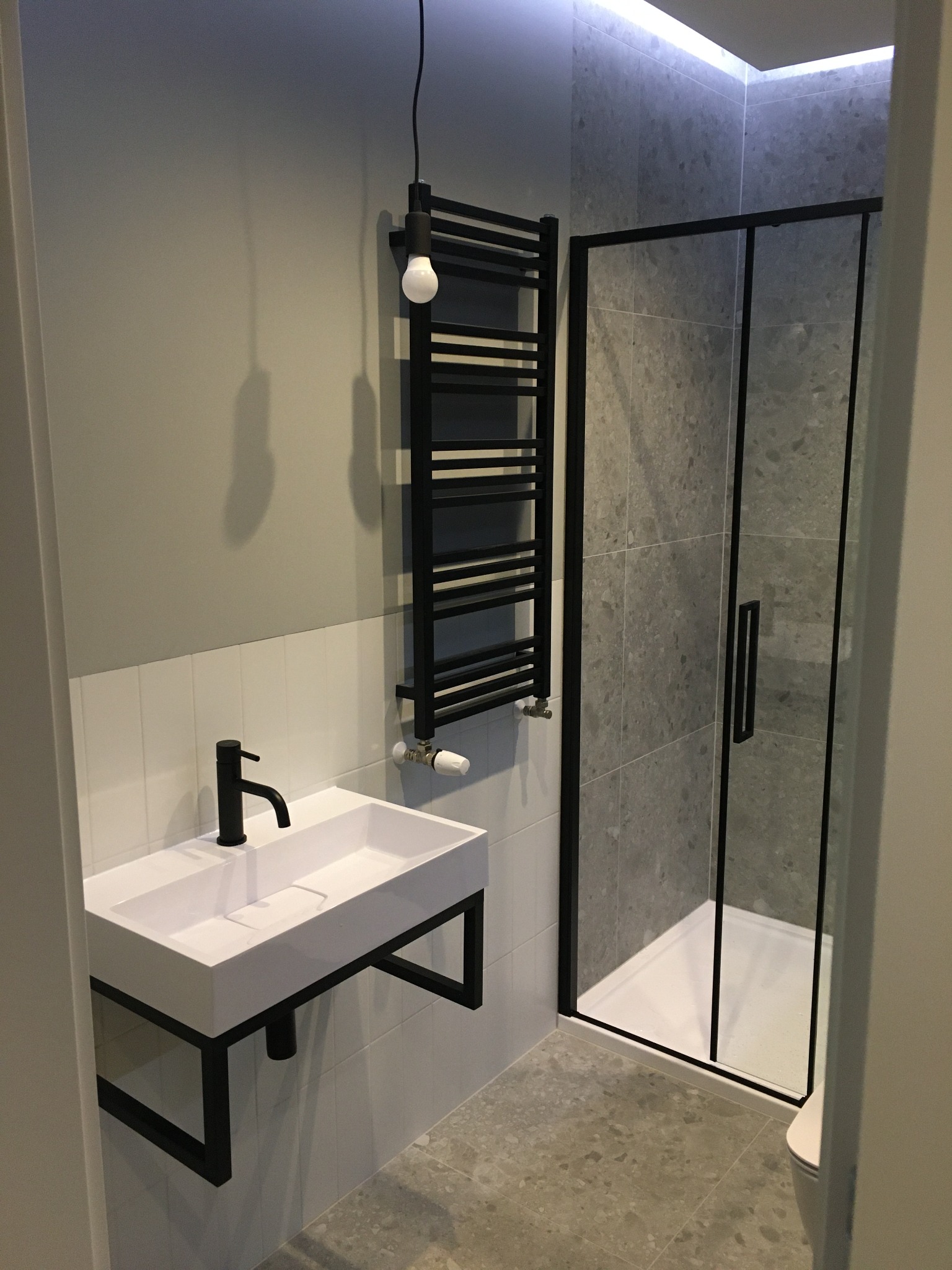 Nowoczesna łazienka z białą umywalką na czarnym stelażu, czarną armaturą, grzejnikiem drabinkowym i kabiną prysznicową z szarymi płytkami imitującymi kamień.