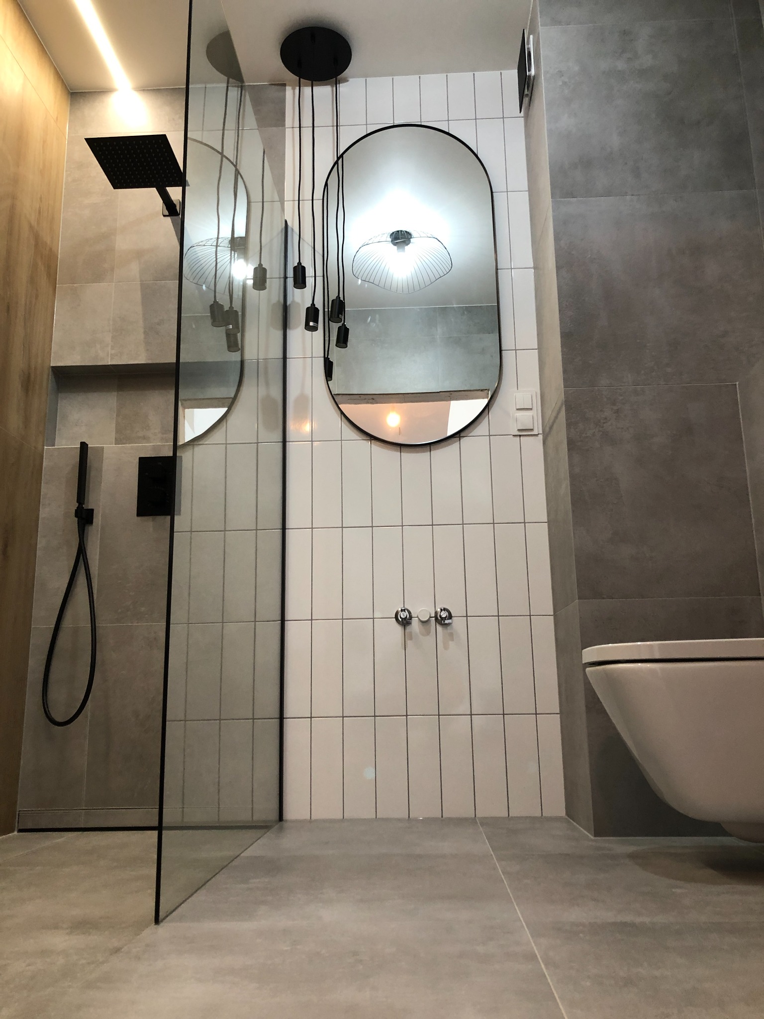 Nowoczesna łazienka z kabiną prysznicową typu walk-in, szarymi i białymi płytkami, minimalistycznym oświetleniem i owalnym lustrem.